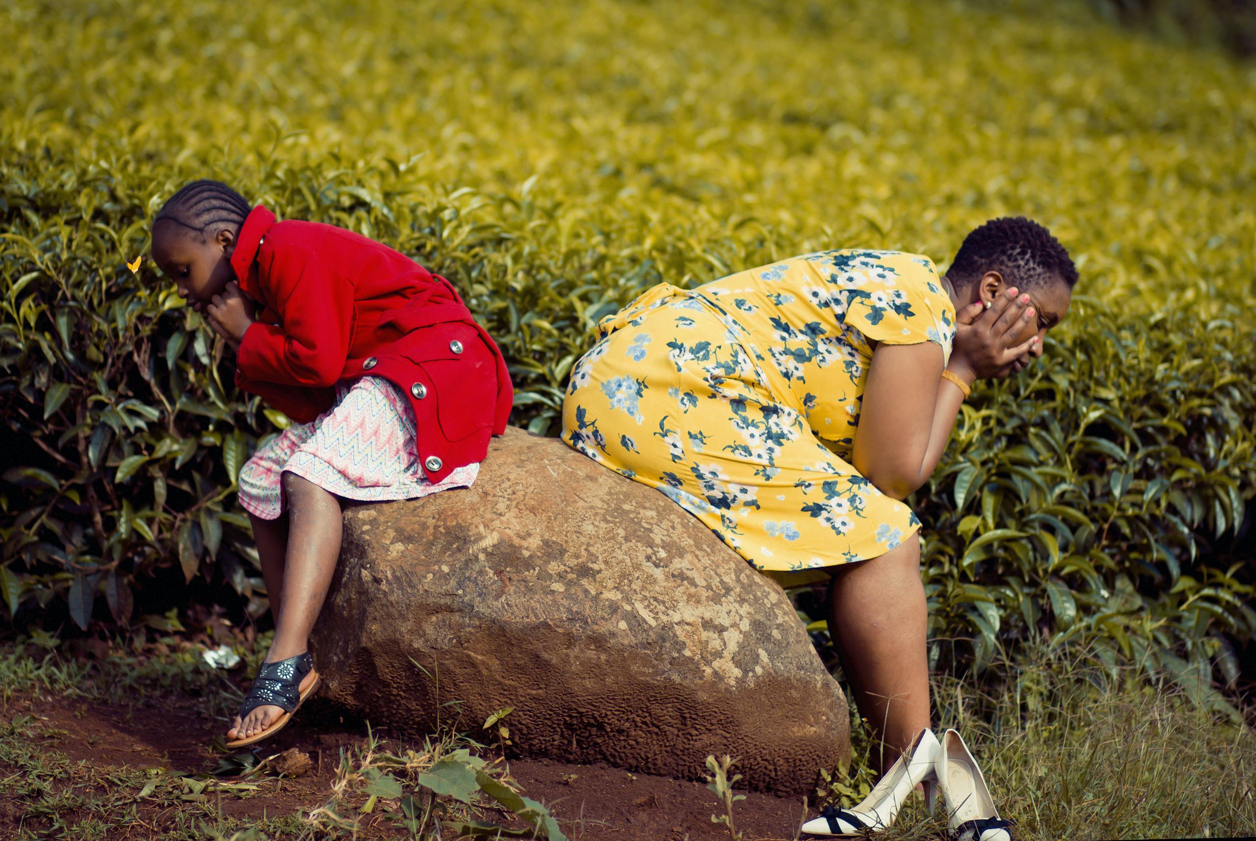 A woman and child seated back-to-back on a rock in a lush green field, deep in thought.